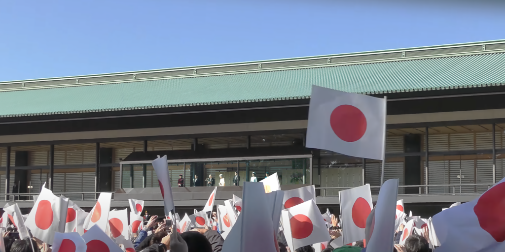 A crowd for Emperor Akihito's 84th birthday at the Imperial Palace in Tokyo. The crowd waves small Japanese flags in the foreground and the imperial family stand on the balcony in the background.