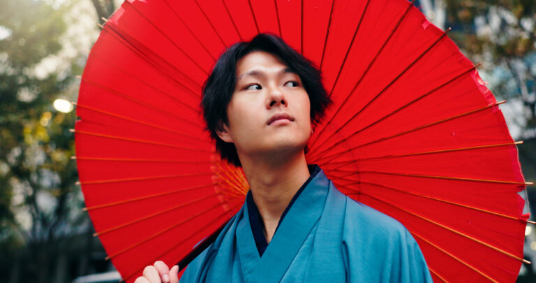 A handsome (ikemen) Japanese man in a blue traditional garment stands holding a large red parasol, looking to the side. Trees and buildings are visible in the blurred background.