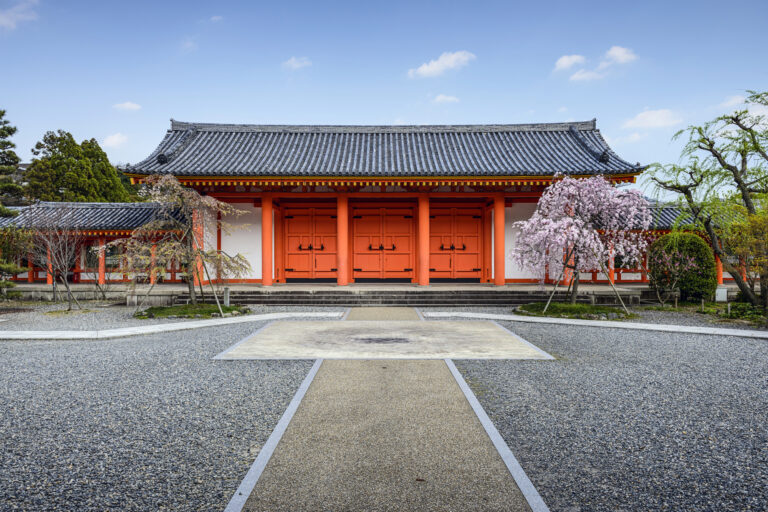 Traditional Japanese temple gate with red doors, surrounded by gravel paths and blooming cherry blossoms, embodies the serene Japanese concepts and philosophies found in Zen Buddhism and Japanese culture