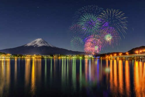 Fireworks light up the night sky above a lake, their reflections shimmering on the water with a snow-capped mount Fuji and sparkling city lights in the background.