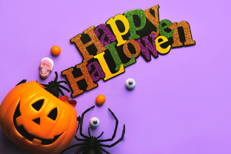 A plastic jack-o'-lantern, fake spiders, eyeball candies, skull, and a "Happy Halloween" sign with happy halloween in Japanese are arranged on a purple background.