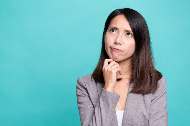 A woman in a gray blazer looks up thoughtfully with her hand on her chin, as if pondering japanese question words, standing in front of a plain turquoise background.