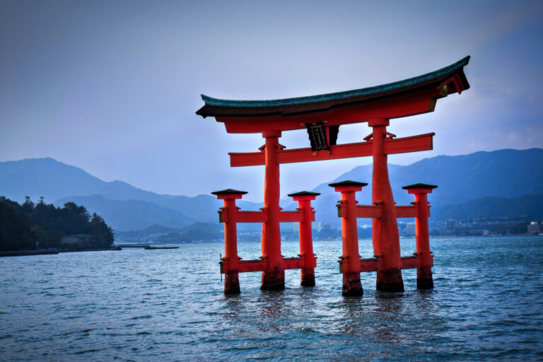 A large red Japanese torii gate stands in the water with mountains and a town visible in the background under a cloudy sky.