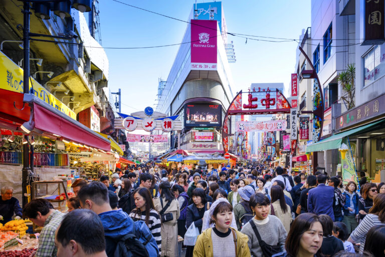 A busy outdoor market scene in Tokyo with a large crowd of shoppers, colorful stalls, and signs in Japanese all set under a bright blue sky.