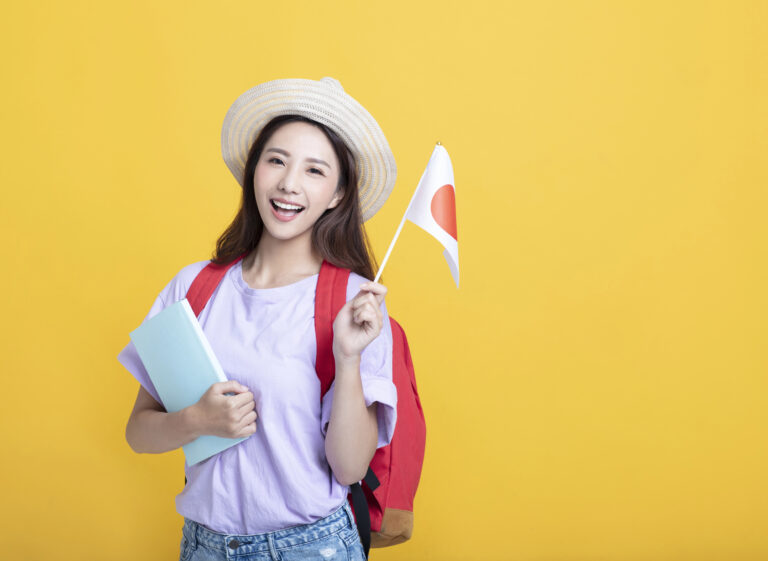 A young woman with a backpack holds notebooks and a small Japanese flag, smiling in front of a yellow background