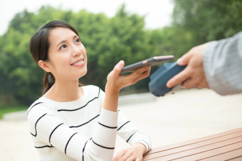 A woman sitting at an outdoor table pays with her smartphone using a contactless terminal, then smiles and says "gochisousama," expressing gratitude for the meal.