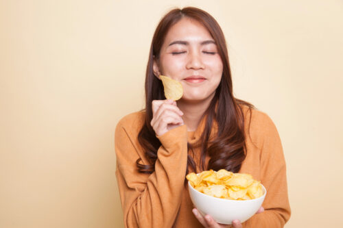A woman in an orange sweater, looking "onaka suita" (hungry in Japanese), holds a bowl of potato chips and a single chip, eyes closed and smiling softly as she stands against a beige background.