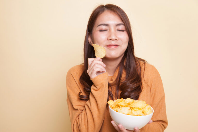 A woman in an orange sweater, looking "onaka suita" (hungry in Japanese), holds a bowl of potato chips and a single chip, eyes closed and smiling softly as she stands against a beige background.