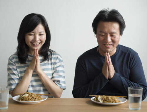 A woman and a man sit at a table with plates of noodles, hands clasped in gratitude as they say "itadakimasu" in Japanese before eating. Two glasses of water are on the table, ready for their meal.