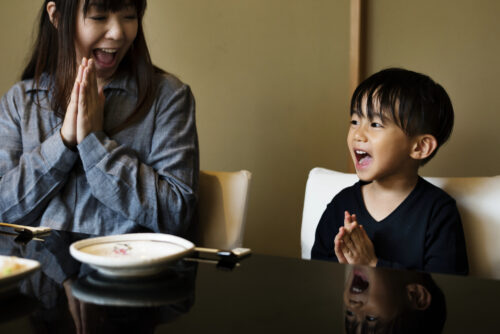 A woman and a young boy sit at a table, smiling and pressing their hands together as they say "itadakimasu" (thanks for the food in Japanese) before enjoying their meal.