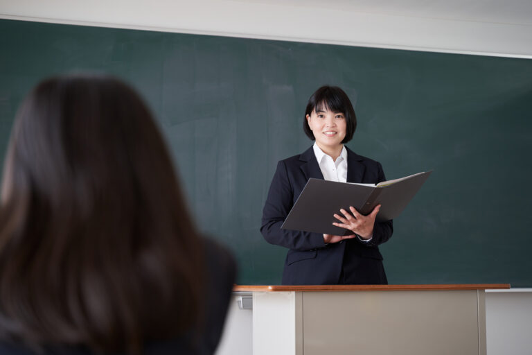 A Japanese female teacher stands at a podium in front of a chalkboard, holding an open binder and speaking to a seated unseen student in a classroom setting.