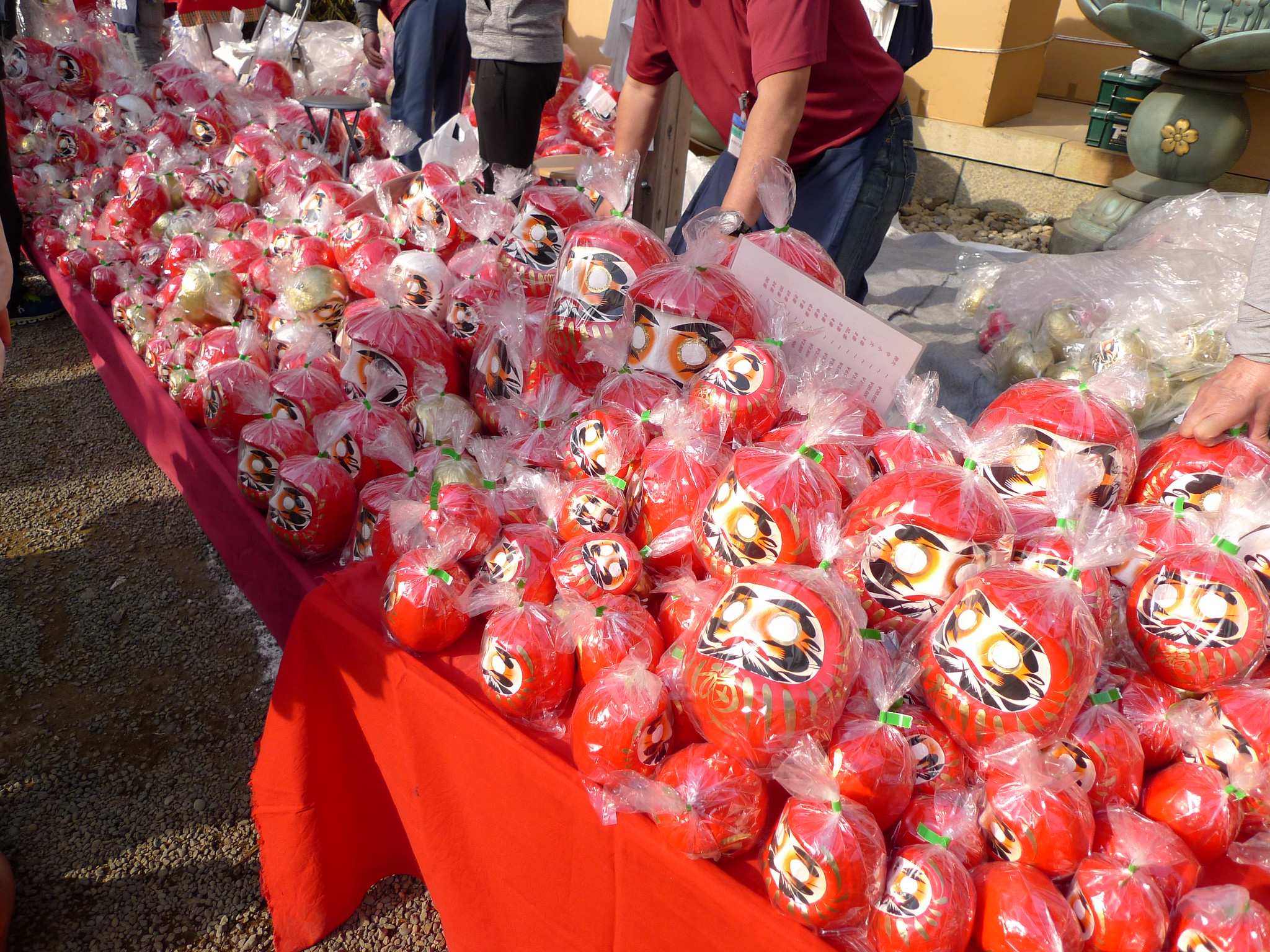 Rows of red Daruma dolls wrapped in plastic bags are displayed on a table with a red cloth at an outdoor 'daruma ichi' market, one of several such markets held around Japan in January.