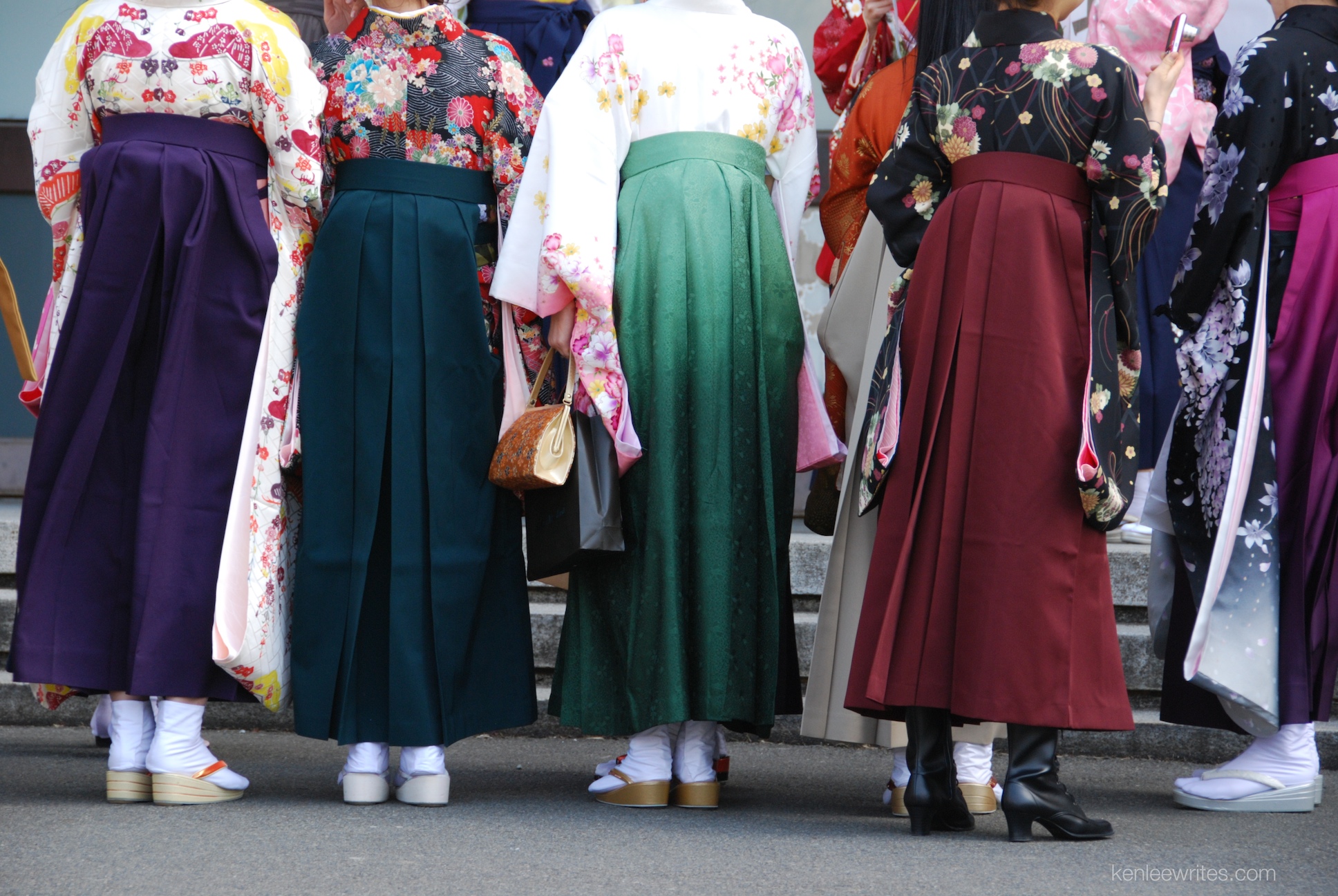 A group of people wearing colorful traditional Japanese hakama and kimono stand in a row, facing away from the camera, as if ready to join a festive march in Japan.