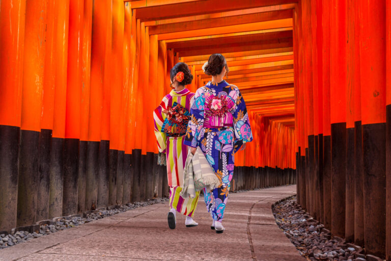 Two women in colorful kimonos walk through a pathway lined with bright orange torii gates.