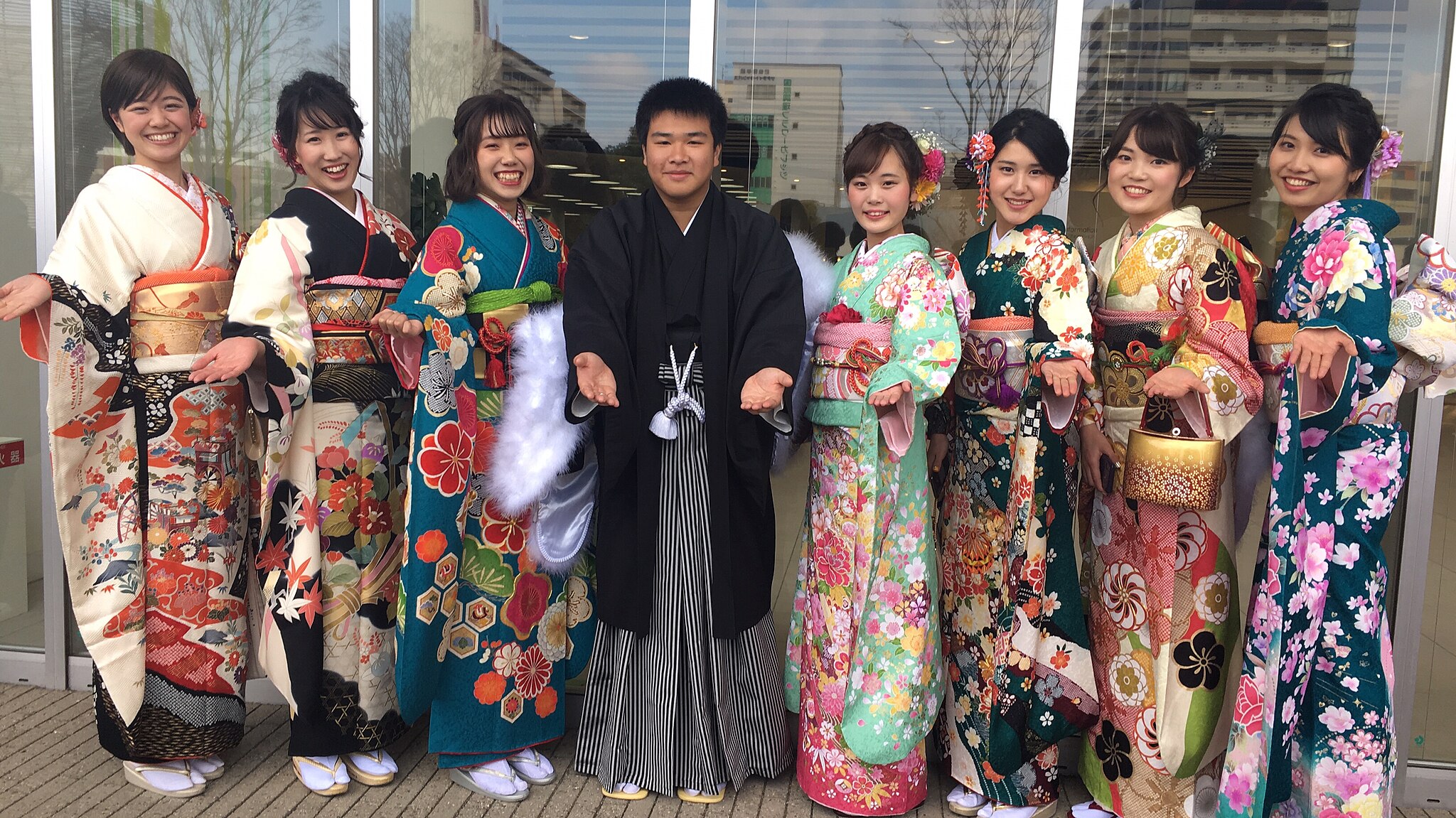 A group of seven women in colorful fursisode (long sleeved kimonos) and one man in traditional Japanese attire (haori and hakama) pose and smile outside a building, holding their hands out, on Coming of Age Day, a national holiday in Japan in January.