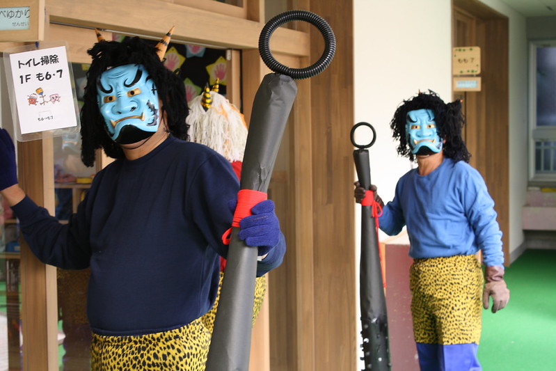 Two people wearing blue ogre masks, blue shirts, and yellow patterned pants stand indoors with large club-like objects, ready to take part in the Setsubun celebrations of February in Japan.