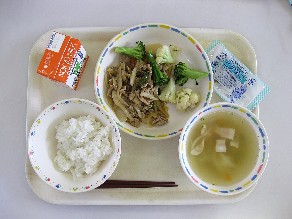 A Japanese school lunch tray with white rice, vegetable and meat stir-fry, miso soup with tofu, a carton of milk, and a packaged fish snack.