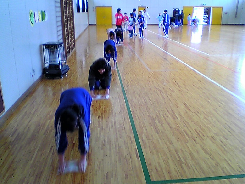 A group of Japanese school children in a gymnasium crawl on the floor in a line, cleaning with cloths, demonstrating how Japanese children clean their own schools daily.