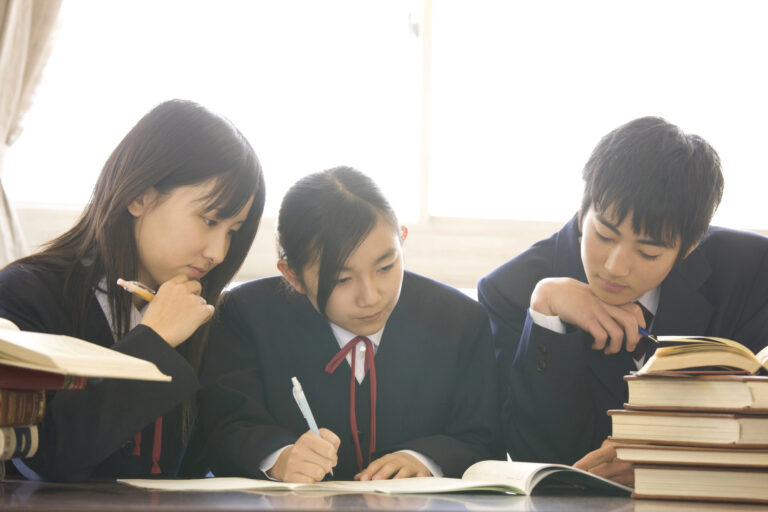 Three Japanese high school students in school uniforms sit at a desk, writing and reading books, with a stack of books nearby and sunlight streaming through the window.