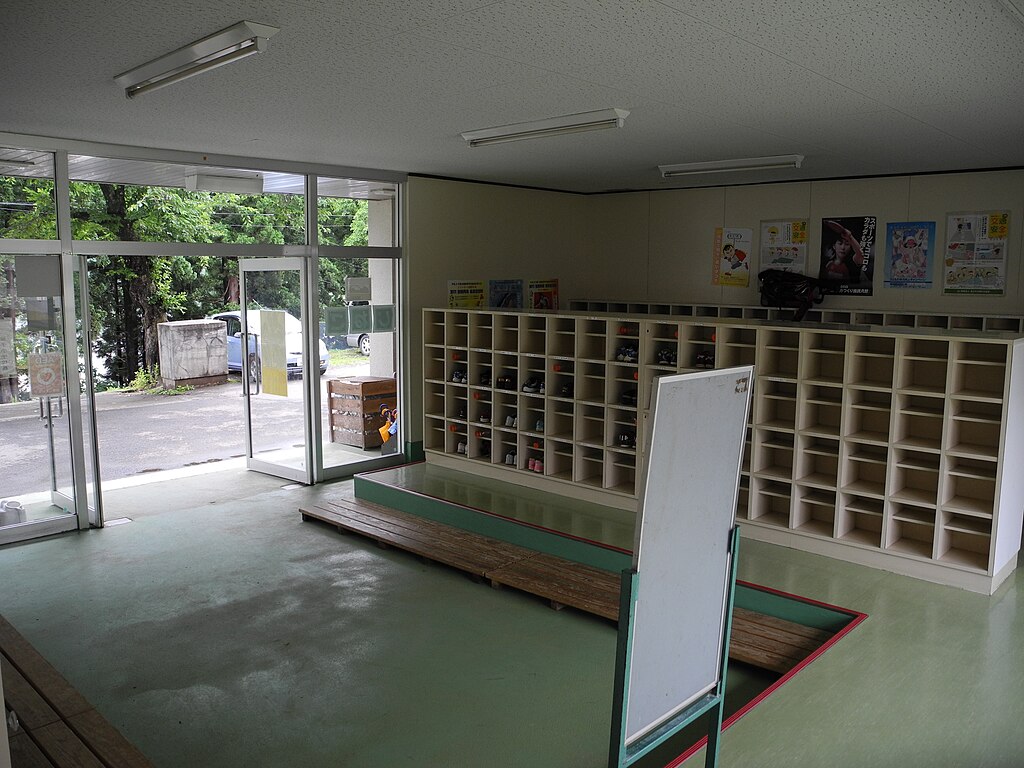 A typical Japanese school entryway with shoe cubbies, a whiteboard, and large open doors showing a view of greenery outside.