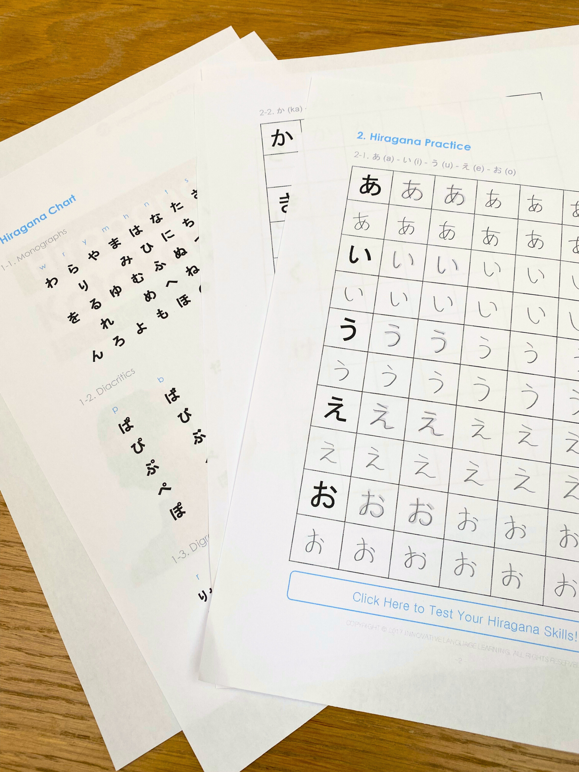 Some printed out hiragana practice sheets for beginners spread on a wooden table, featuring Japanese hiragana charts and practice grids filled with repeated handwritten characters.