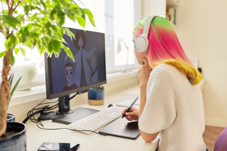 A person with colorful hair and headphones uses a drawing tablet at a desk, with an anime style illustration displayed on a computer monitor.