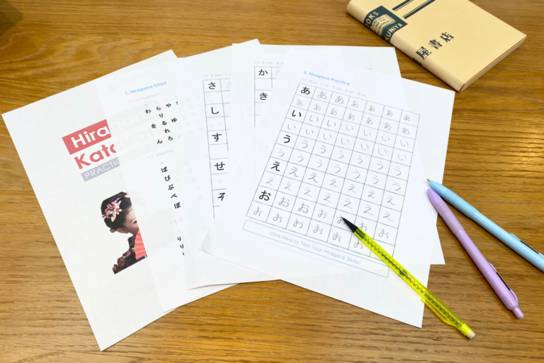 Several Free Hiragana sheets, a book, and three pens are arranged on a wooden desk - tools for beginners to start learning Hiragana.