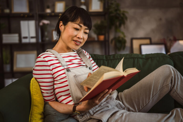 A Japanese woman in a whimsical striped shirt and overalls sits on a couch, reading a hardback book, smiling, with bookshelves and plants in the background. Overall cozy Ghibli vibes.