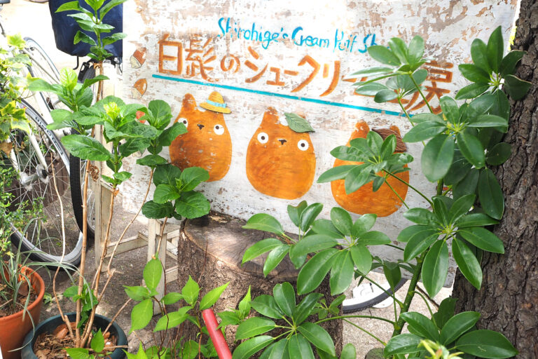 A weathered sign with three orange cartoon characters, some wearing hats and reminiscent of Totoro from the Ghibli movies set in Japan. The sign is surrounded by green plants, with a bicycle partially visible in the background.