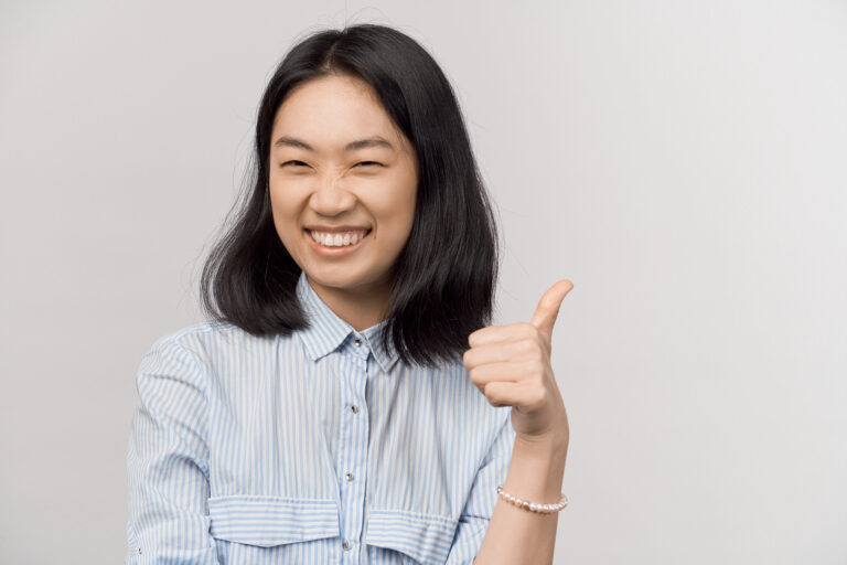 Asian woman with shoulder-length dark hair, wearing a light blue striped shirt, smiles and gives a thumbs-up gesture against a plain light background, as if saying "good job" in Japanese.