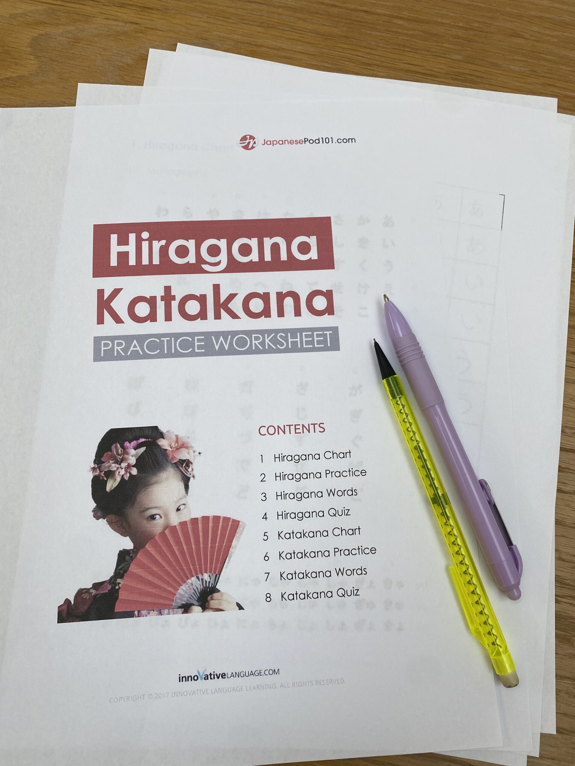 A Hiragana and Katakana practice worksheet with a girl holding a fan on the cover, beside a purple pen and a yellow mechanical pencil on a wooden table - download these Japanese practice sheets for free here.