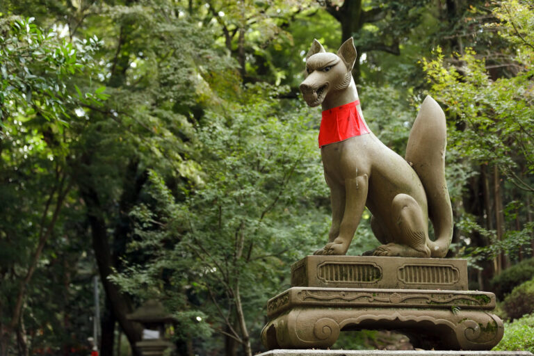 A stone fox statue with a red bib sits on a pedestal surrounded by lush green trees, evoking the spirit of Japanese traditions seen in Ghibli movies.
