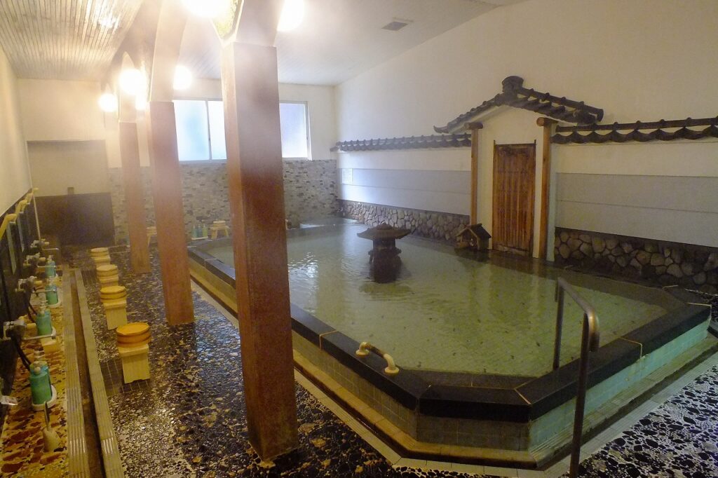 Indoor Japanese onsen bath with wooden stools and washing stations lining the perimeter, warm lighting, and a central fountain in the bathing pool, capturing the serene atmosphere often seen in Ghibli movies and rooted in Japanese traditions.