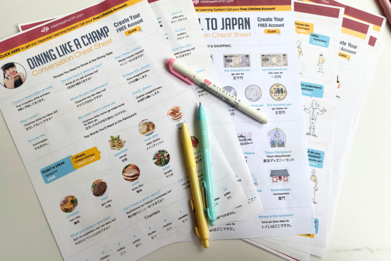 Several Japanese language cheat sheets and guides with food images and illustrations are spread out on a desk, a selection of free Japanese printables, with three pastel-colored pens and a pink mechanical pencil placed on top.