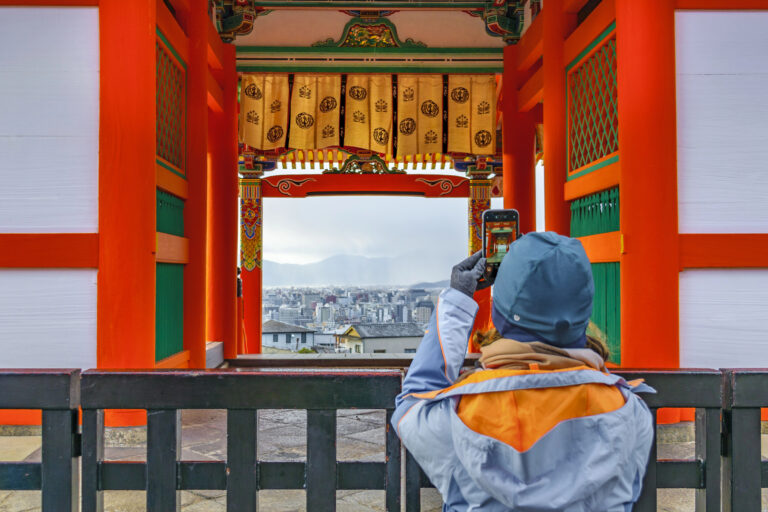 Person in a blue jacket and hat takes a photo of a cityscape through the ornate red and yellow entrance gate of a traditional Japanese temple.