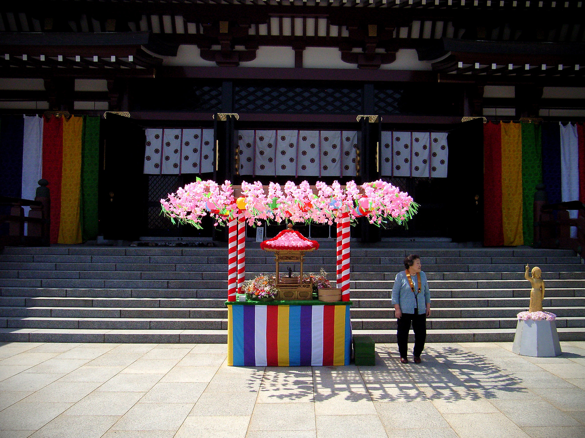 A woman stands beside a colorful, flower-decorated booth in front of temple steps during hanamatsuri in April in Japan, with banners and a small golden statue nearby.