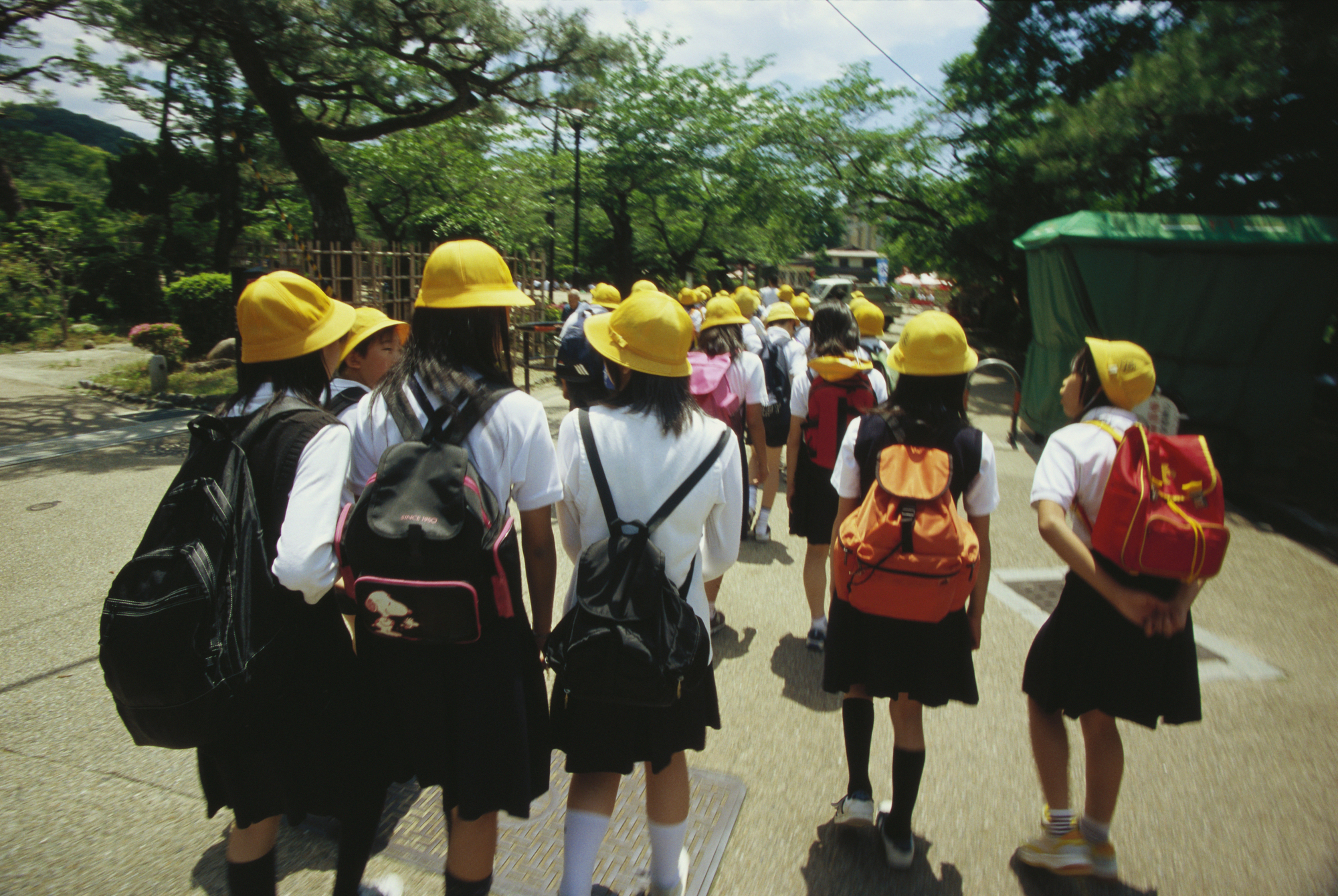 A group of schoolchildren wearing yellow hats and uniforms walk outdoors in a line, carrying backpacks, with trees and a clear sky in the background—a typical scene during April in Japan, which is the start of the new school year.