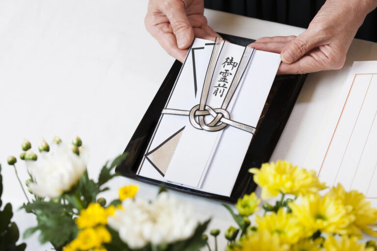 Hands holding a traditional Japanese envelope on a tray, with yellow and white flowers in the foreground.