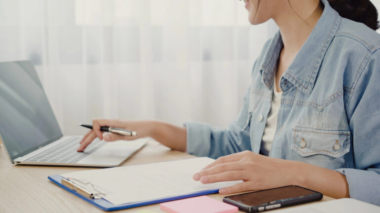 Person in a denim jacket studying Japanese at a desk with a laptop, clipboard, notepad, and smartphone.