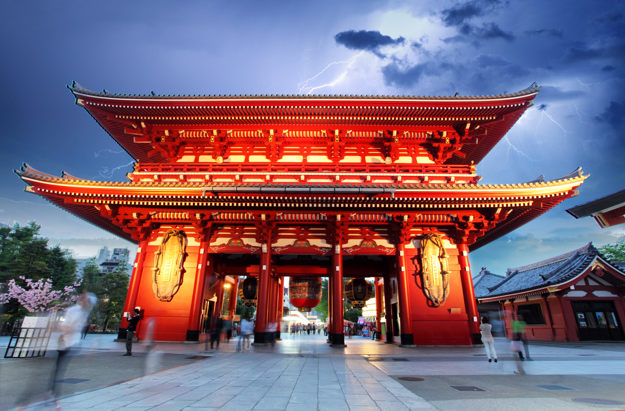 The red entrance gate of a traditional Japanese temple, illuminated at dusk with blurred people passing and lightning streaks in a stormy sky above.