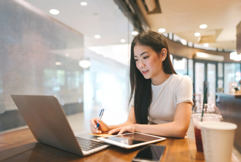 A young Asian woman sits at a table with a laptop, tablet, smartphone, and drinks, writing in her notebook—perhaps studying Japanese with japanesepod101 premium plus — in a bright, modern cafe.