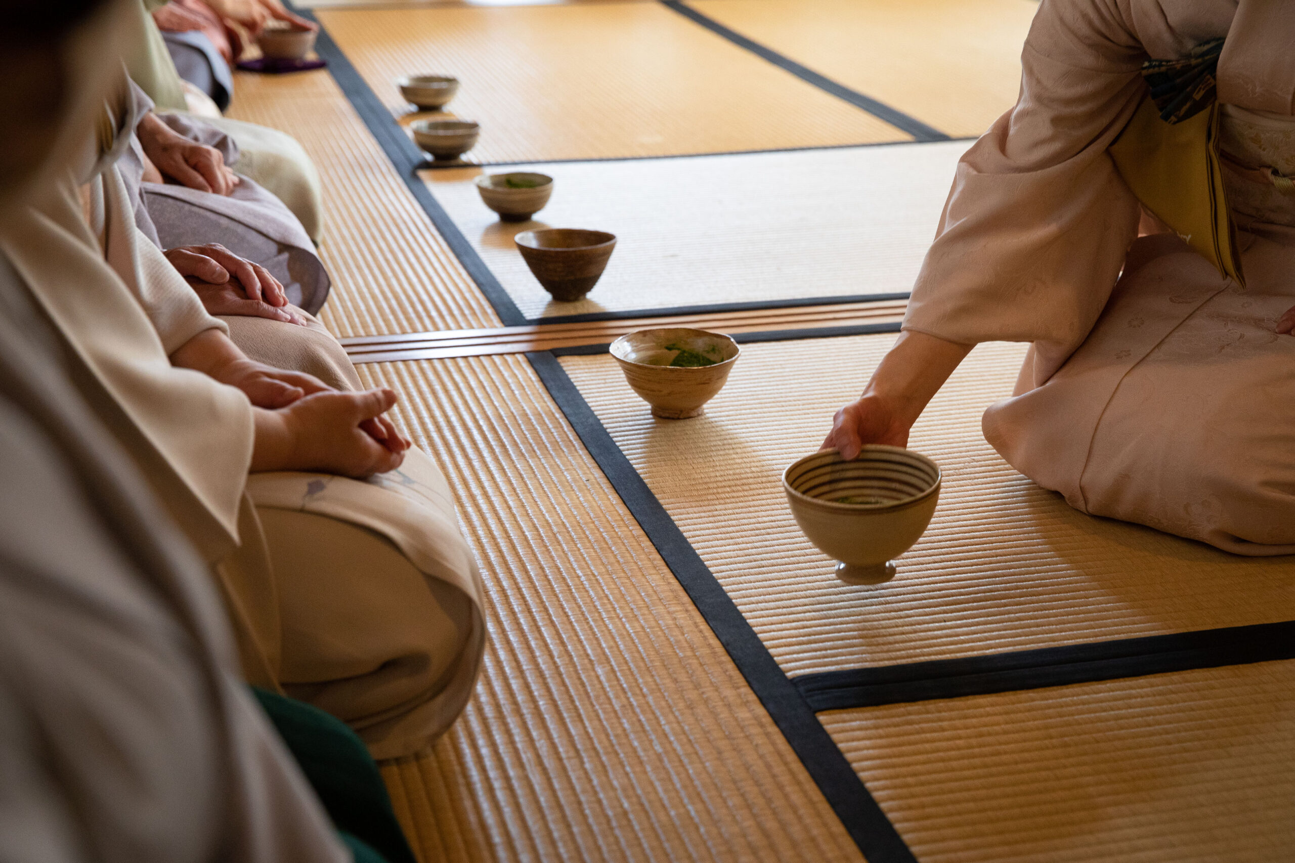 People in traditional clothing participate in a Japanese Tea Ceremony, sitting on tatami mats with bowls placed in front of them, carefully observing tea ceremony etiquette.