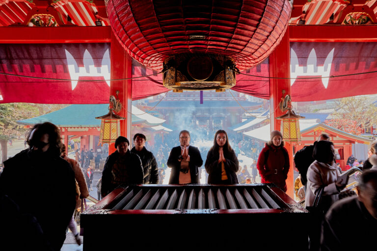 People stand and pray at a large red shrine in Japan, with incense smoke drifting in the background.