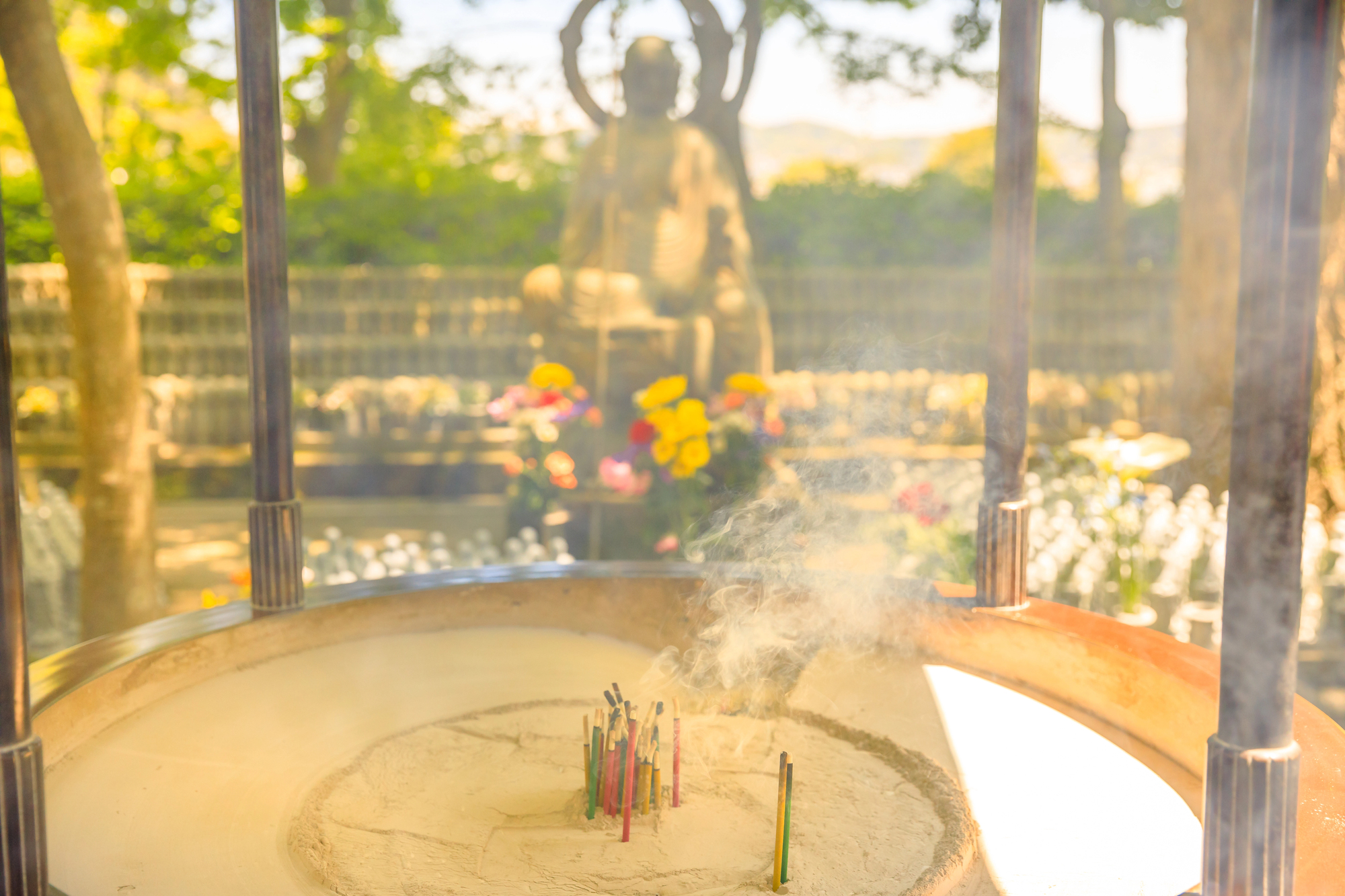 A large incense burner with multiple sticks smoking in front of a blurred Buddha statue and colorful flowers, typical of a Japanese Buddhist temple.