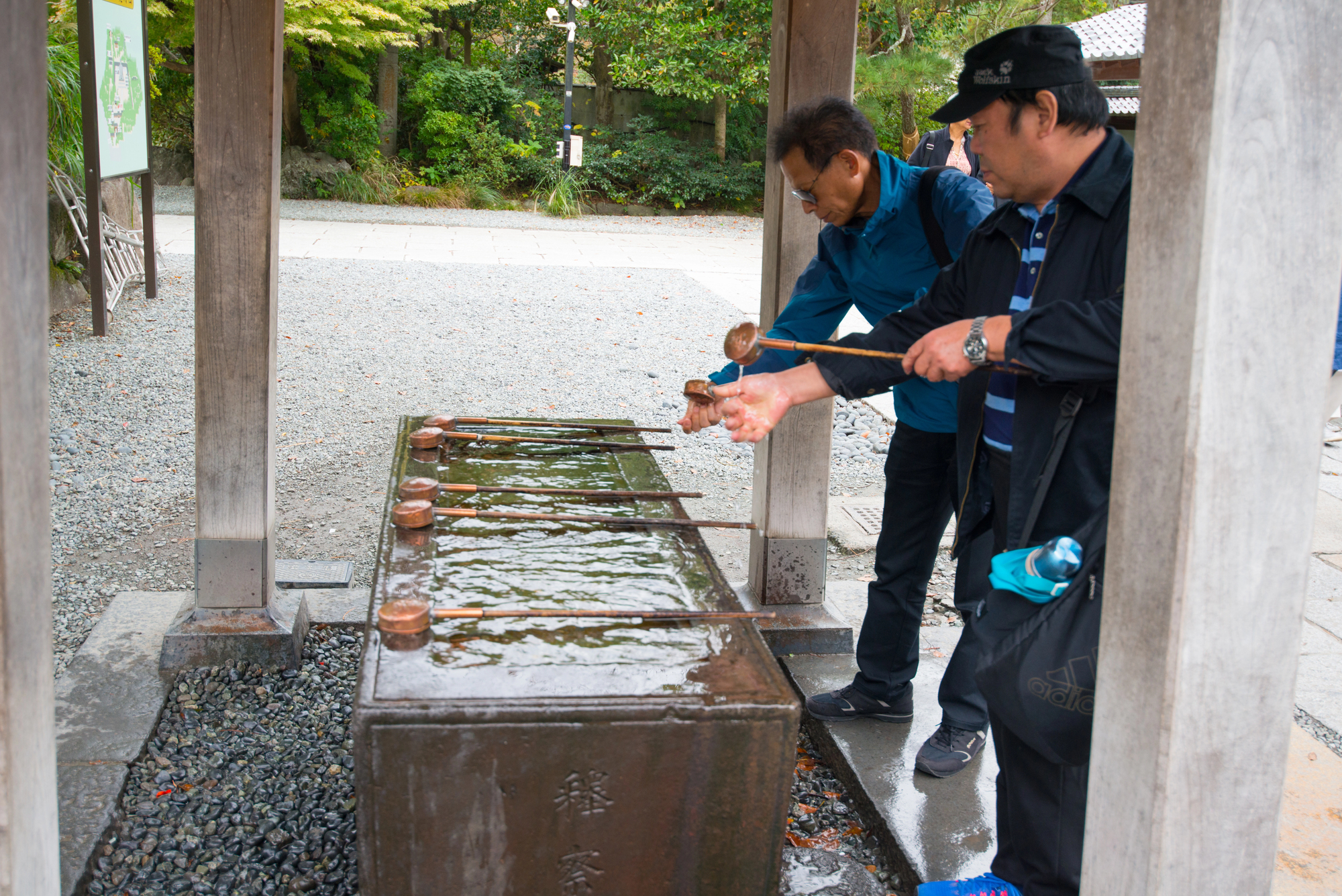 Two men use ladles to wash their hands at a traditional purification fountain, demonstrating proper shrine etiquette in Japan.
