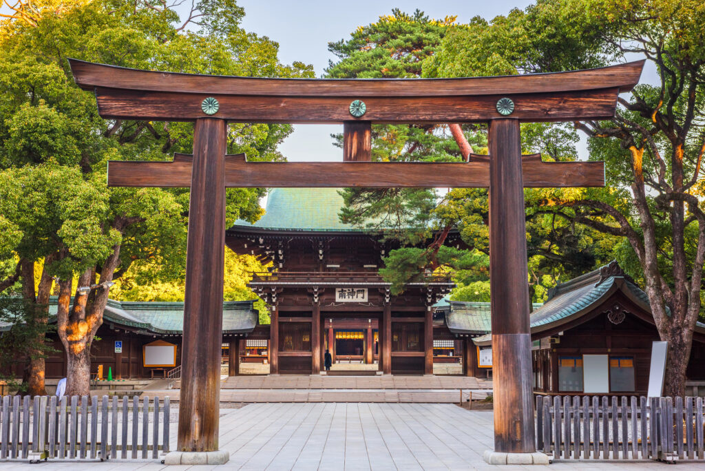 A large wooden torii gate stands in front of a traditional Japanese shrine building, surrounded by green trees and a paved courtyard—an inviting scene for anyone interested in Japan travel or learning about shrine etiquette.