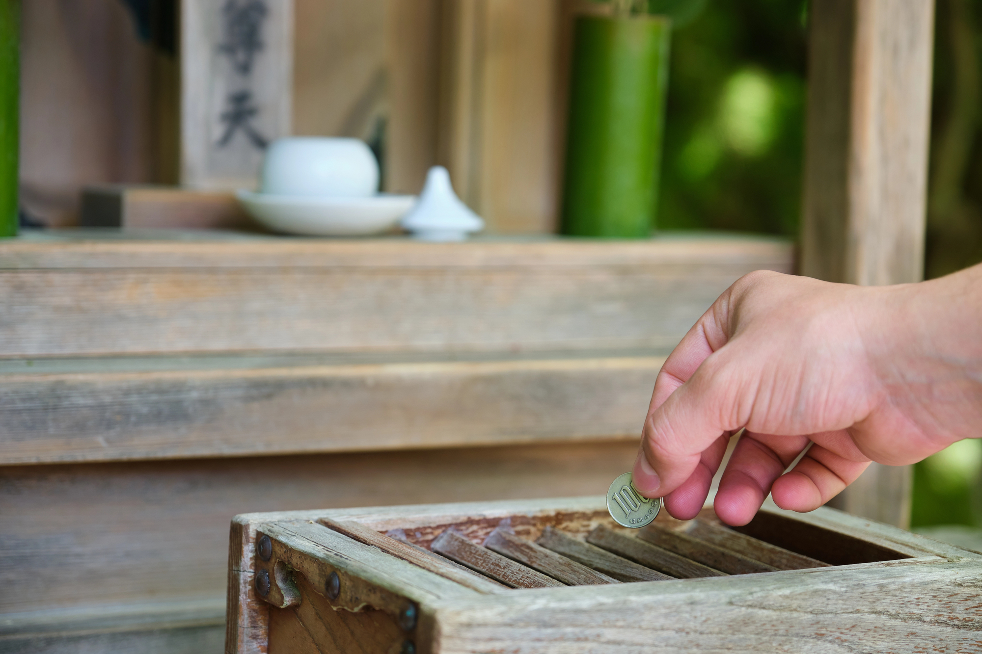 A hand drops a 100 yen coin into a wooden offering box at a Japanese shrine, with a small altar and ritual items visible in the background.