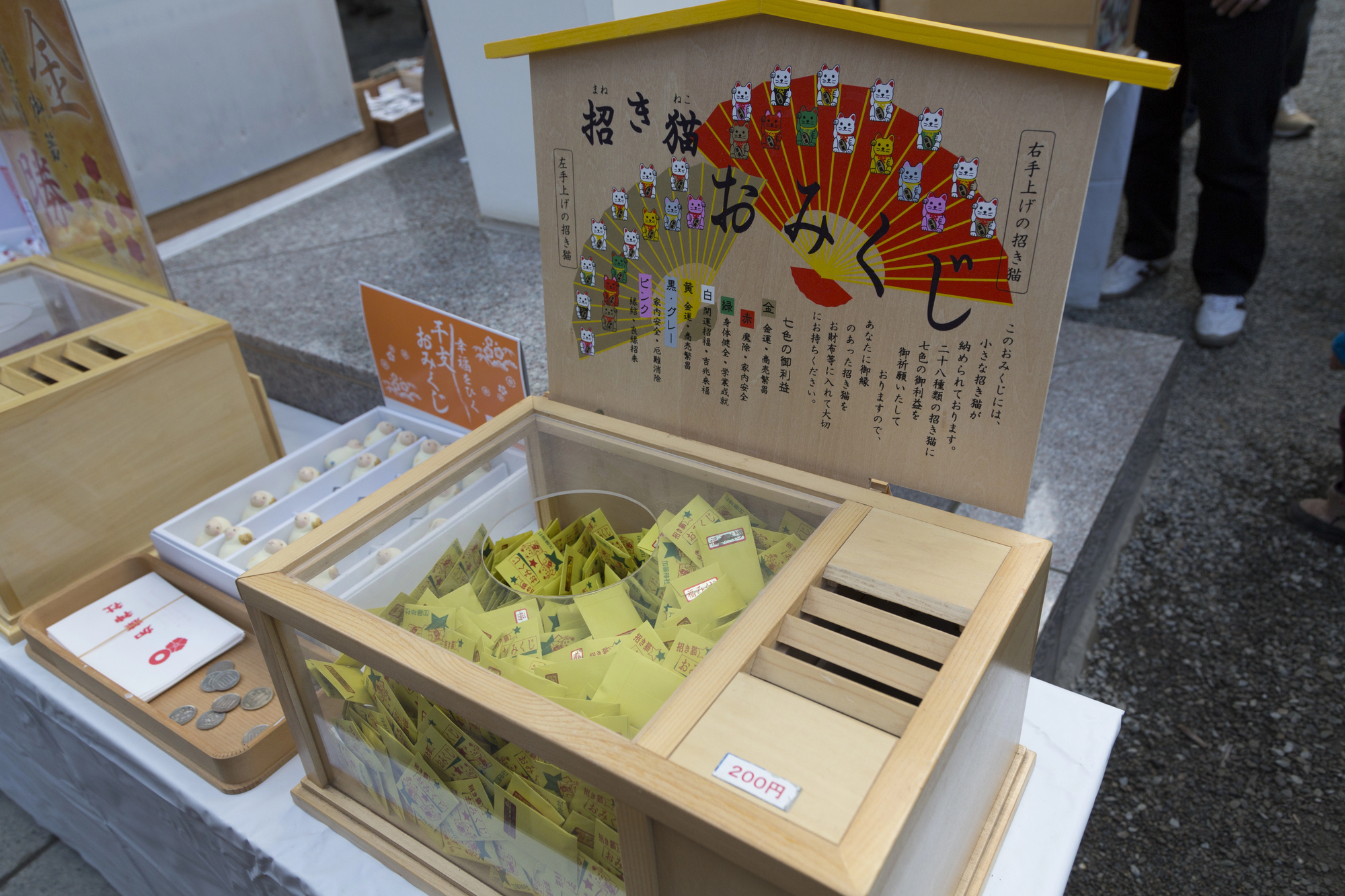 A wooden omikuji fortune box with yellow paper fortunes and cartoon images is displayed on a table beside a payment slot labeled "200円" and scattered coins at a temple shrine in Japan.