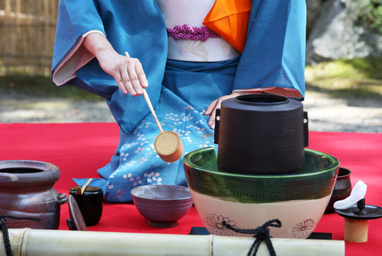 Person in traditional Japanese attire prepares matcha using a bamboo ladle at an outdoor Japanese Tea Ceremony, with tea utensils arranged on a red mat, reflecting the grace and precision of tea ceremony etiquette.