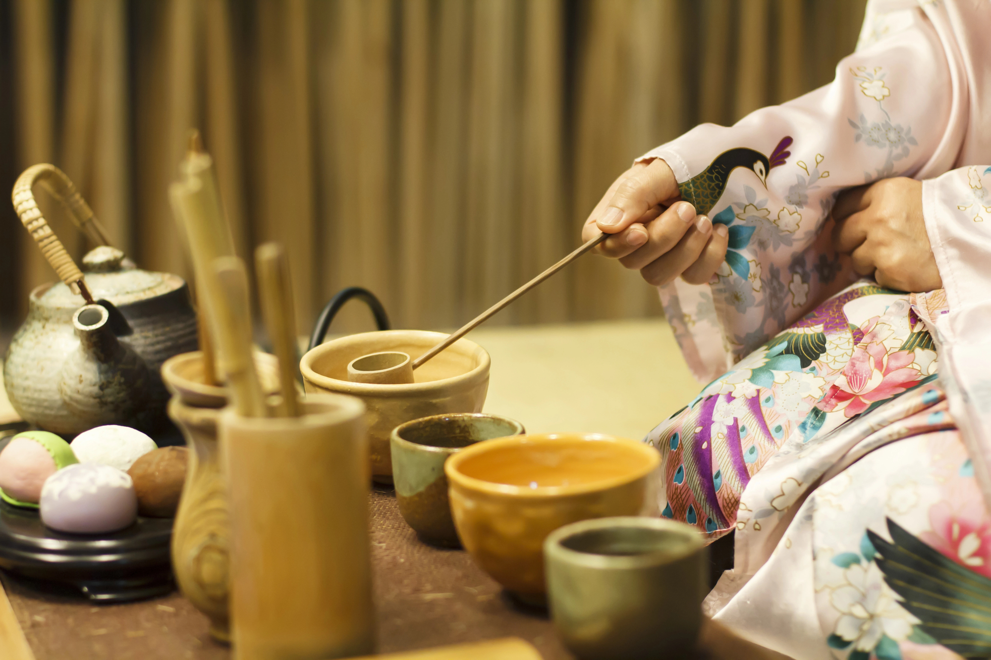 Person in a floral kimono prepares tea using traditional Japanese utensils, embracing Japanese tea ceremony etiquette and Japanese tea vocabulary, with sweets on the side for an authentic experience.
